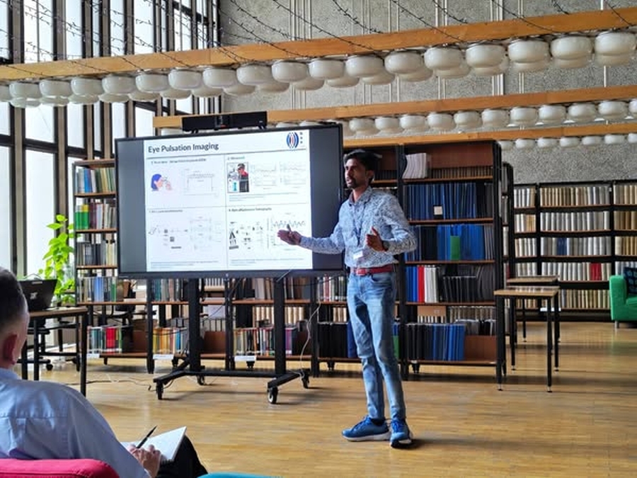 PhD student Vasantha Kumar Kathirvelu gives a presentation on eye pulsation imaging in a modern library setting, standing beside a screen with diagrams and graphs, while an audience member takes notes