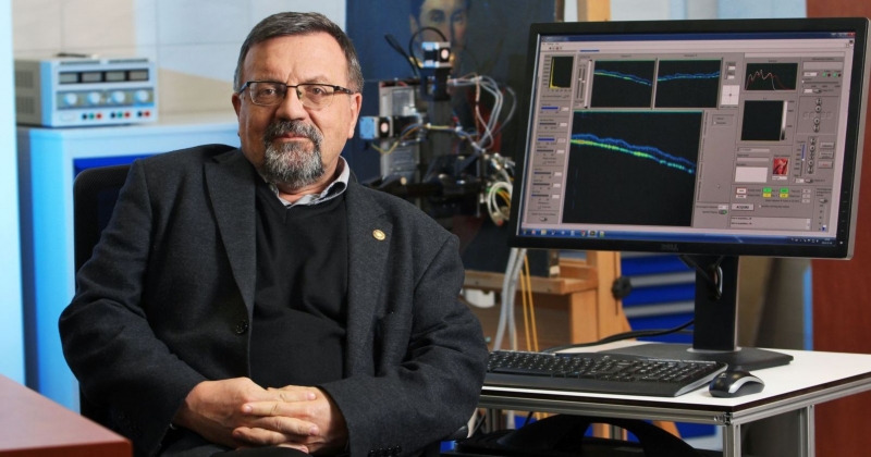 Zdjęcie portretowe prof. Piotra Targowskiego. Na monitorze komputera widać oprogramowanie specjalistyczne, a w tle stoi także sprzęt do tomografii optycznej analizujący obraz na sztaludze