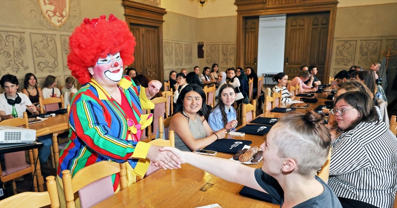 Osoba przebrana za clowna podaje rękę jednemu z uczestników kursu. Wokół przy stole siedzą inni kursanci