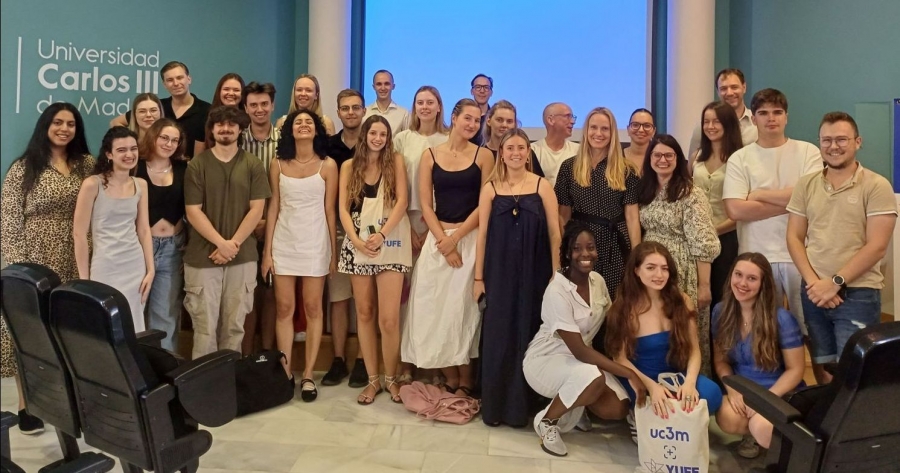 About 30 young smiling people pose for a group photo. On the wall to the left is the inscription ?Universidad Carlos III de Madrid.?