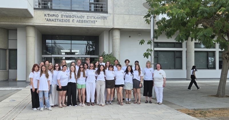 Na pierwszym planie grupa uśmiechniętych studentów oraz nauczycieli stojących w szeregu na tle wejścia do budynku Uniwersytetu Cypryjskiego.