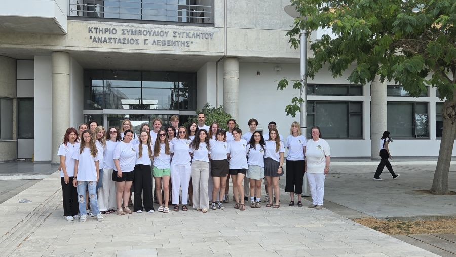 In the foreground, a group of smiling students and teachers stand in a row against the the entrance to the University of Cyprus building.