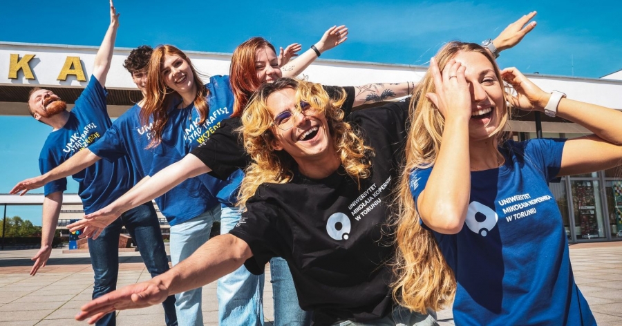 Grupa uśmiechniętych studentów i studentek UMK z rozłożonymi ramionami. Ubrani są w koszulki z logo UMK lub napisem 