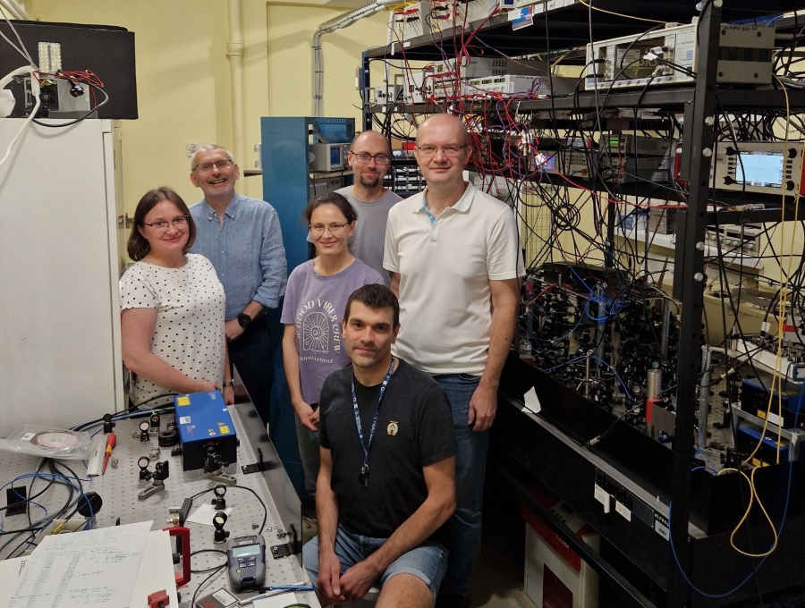 Grupa sześciu osób (dr hab Katarzyna Bielska, prof. UMK, prof. dr hab. Roman Ciuryło, dr hab. Agata Cygan, prof. UMK, dr Szymon Wójtewicz, prof. dr hab. Daniel Lisak i prof. dr hab. Piotr Wcisło) w laboratorium naukowym. Wszyscy pozują do zdjęcia. Profesor Piotr Wcisło siedzi na pierwszym planie, a pozostałe pięć osób stoi za nim. Ubrani są w swobodny, codzienny strój. Otoczenie jest wypełnione specjalistycznym sprzętem. Po lewej stronie widoczny jest stół optyczny z różnymi komponentami, w tym z niebieskim urządzeniem, prawdopodobnie laserem. Po prawej stronie znajduje się wysoka szafa rackowa, gęsto wypełniona aparaturą elektroniczną i połączona licznymi kablami. Tło również jest zapełnione aparaturą badawczą.