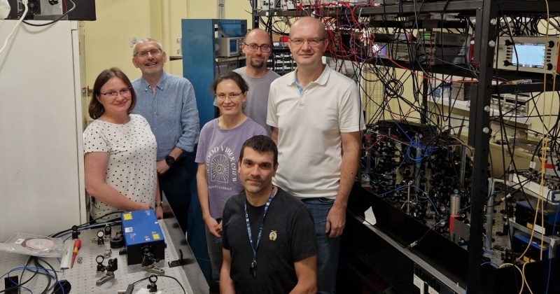 Grupa sześciu osób (dr hab Katarzyna Bielska, prof. UMK, prof. dr hab. Roman Ciuryło, dr hab. Agata Cygan, prof. UMK, dr Szymon Wójtewicz, prof. dr hab. Daniel Lisak i prof. dr hab. Piotr Wcisło) w laboratorium naukowym. Wszyscy pozują do zdjęcia. Profesor Piotr Wcisło siedzi na pierwszym planie, a pozostałe pięć osób stoi za nim. Ubrani są w swobodny, codzienny strój. Otoczenie jest wypełnione specjalistycznym sprzętem. Po lewej stronie widoczny jest stół optyczny z różnymi komponentami, w tym z niebieskim urządzeniem, prawdopodobnie laserem. Po prawej stronie znajduje się wysoka szafa rackowa, gęsto wypełniona aparaturą elektroniczną i połączona licznymi kablami. Tło również jest zapełnione aparaturą badawczą.