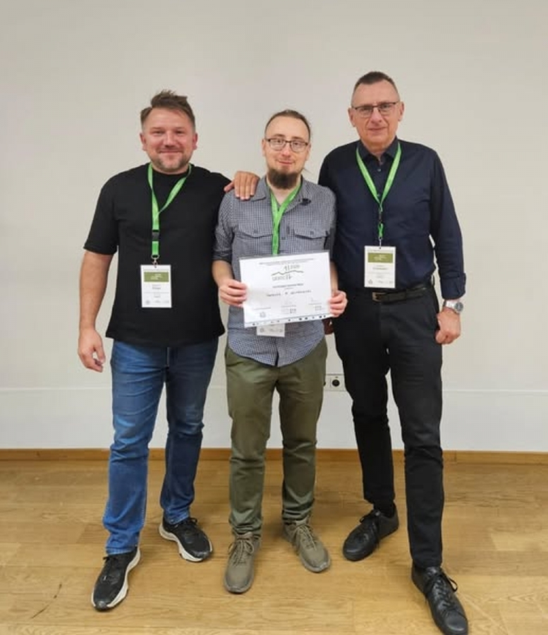 Three men standing side by side at an event; the man in the middle is holding a certificate, and all are wearing name badges with green lanyards, smiling for the photo.
