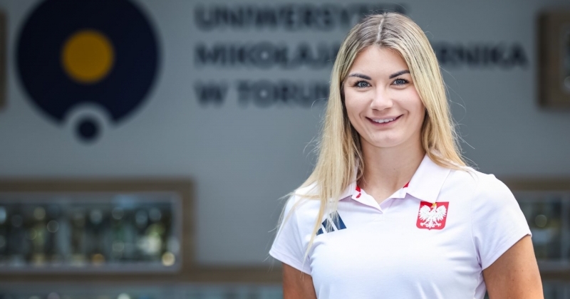 Na zdjęciu znajduje się młoda kobieta z blond włosami, ubrana w białą koszulkę polo z godłem Polski i logo Adidas. Stoi na tle napisu ?Uniwersytet Mikołaja Kopernika w Toruniu? oraz gablot z pucharami. Uśmiecha się do obiektywu.