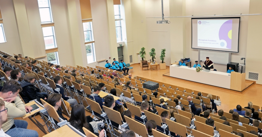 Zdjęcie przedstawia uroczystość akademicką w dużej auli uniwersyteckiej, gdzie studenci i goście siedzą w ławkach, a na podium w towarzystwie władz uczelni przemawia profesor w todze.