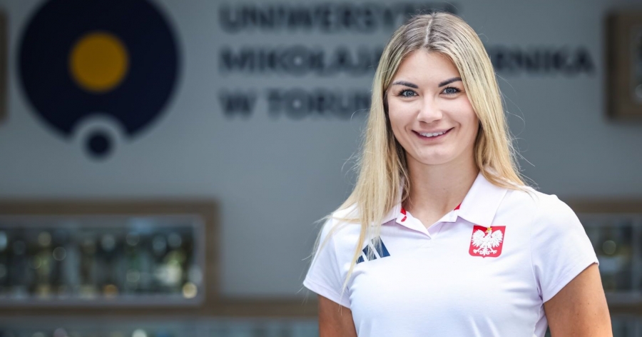 Zdjęcie Anety Rygielskiej w koszulce reprezentacyjnej z orzełkiem na piersi, w tle rozmyte logo UMK