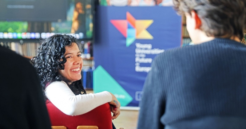 A smiling student turned sideways to the student sitting behind her. In the background, you can see a roll-up banner with the YUFE logo.