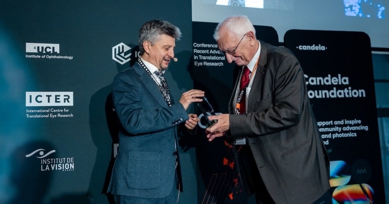 Zdjęcie przedstawia scenę z konferencji naukowej lub medycznej, na której prof. dr hab. Maciej Wojtkowski wręcza prof. dr. hab. Andrzejowi Kowalczykowi statuetkę. Prof. dr hab. M. Wojtkowski stoi przodem do prof. dr. hab. A. Kowalczyka i lekko bokiem do obiektywu. Jest to mężczyzna w średnim wieku, z krótkimi, szpakowatymi włosami. Ubrany jest w elegancki, ciemnogranatowy garnitur i krawat. Na szyi ma zawieszoną smycz identyfikacyjną. Właśnie przekazuje statuetkę do rąk drugiego mężczyzny. Uśmiecha się. Prof. dr hab. A. Kowalczyk jest starszy, ma siwe włosy i okulary. Ubrany jest w ciemny, białą koszulę i czerwony krawat. Na szyi ma również zawieszoną smycz z identyfikatorem. Pochyla się nieco do przodu, by przyjąć nagrodę. Uśmiecha się łagodnie. Obaj mężczyźni trzymają w dłoniach małą, przezroczystą statuetkę, która wygląda na wykonaną ze szkła lub akrylu. Scena rozgrywa się na tle ciemnej, profesjonalnej ściany wystawowej, pełnej logotypów i napisów związanych z konferencją. Zdjęcie ma uroczysty i pozytywny charakter, uwieczniając moment wręczenia prestiżowej nagrody podczas międzynarodowej konferencji naukowej. Obaj mężczyźni wydają się zadowoleni i honorowi.