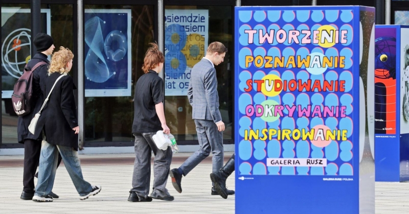 Grupa studentów idzie wzdłuż rektoratu, w centralnym punkcie zdjęcia widać również plakat Galerii Rusz, na której napisano "Tworzenie, poznawanie, studiowanie, odkrywanie, inspirowanie"