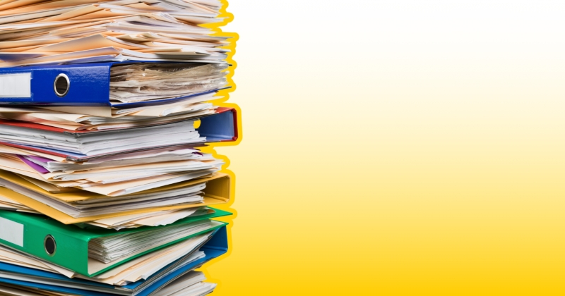 Z lewej storny stos dokumntów: teczki, segregatory i papiery. Żółte tło. 