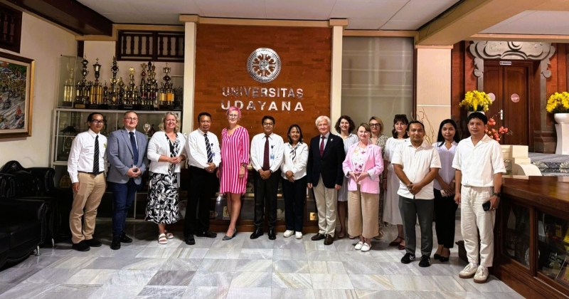 15 osób - delegacja UMK i przedstawiciele Udayana University - stoją w rzędzie. W tle logo indonezyjskiej uczelni