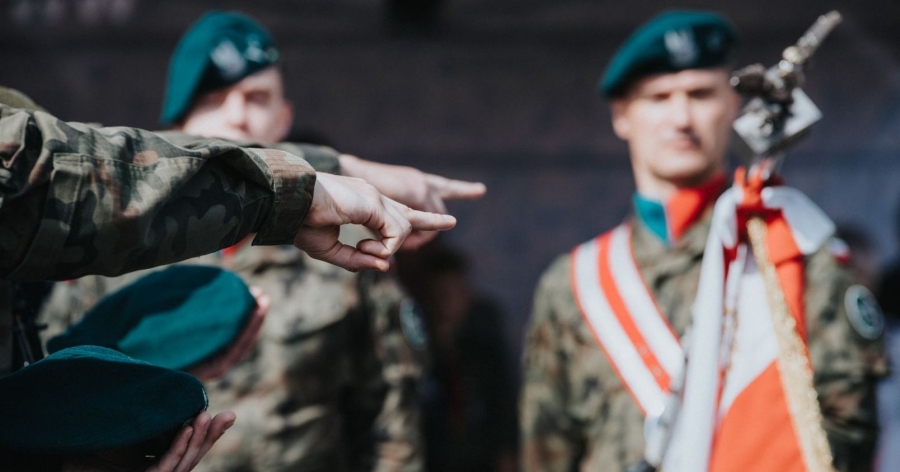 Zdjęcie z przysięgi wojskowej - widać dłonie żołnierzy wskazujące na sztandar, w tle dwóch umundurowanych żołnierzy