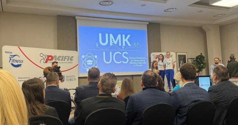 Ludzie w różnych ubraniach siedzą na krzesłach w zamkniętej przestrzeni. Na ekranie wyświetlony jest napis UMK UCS.