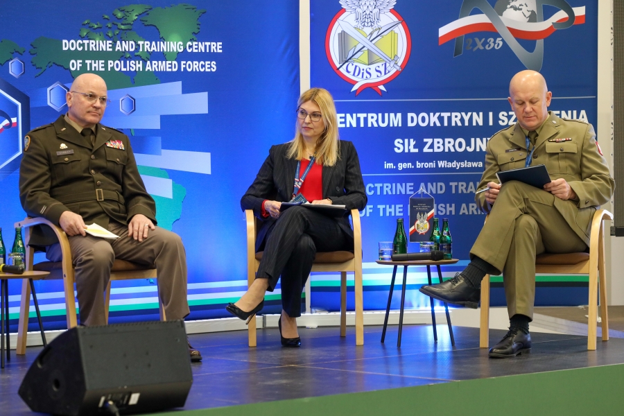 Dwóch mężczyzn w wojskowych mundurach i jedna kobieta ubrana po cywilnemu siedzą na scenie.