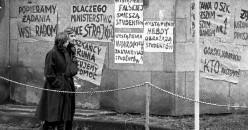 zdjęcie przedstawia dwoje studentów spacerujących na tle muru obklejonego plakatami strajkowymi