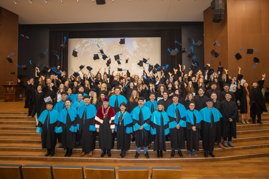 Aula UMK. W pierwszym rzędzie władze dziekańskie i promotorzy w strojach akademickich. Za nimi absolwenci podrzucają birety. 