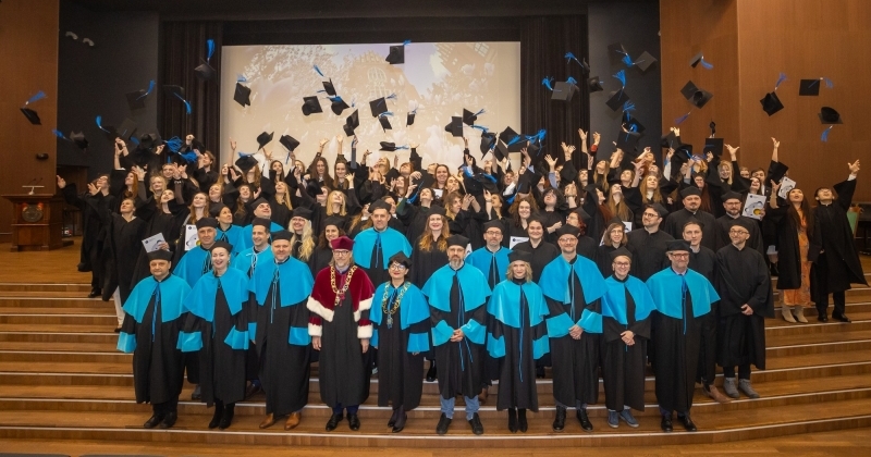 Aula UMK. W pierwszym rzędzie władze dziekańskie i promotorzy w strojach akademickich. Za nimi absolwenci podrzucają birety. 