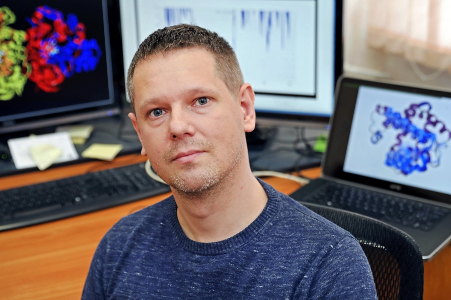 Portret dr. hab. Łukasza Pepłowskiego, prof. UMK, siedzącego przy biurku na tle monitorów wyświetlających trójwymiarowe modele struktur molekularnych oraz wykresy badawcze.