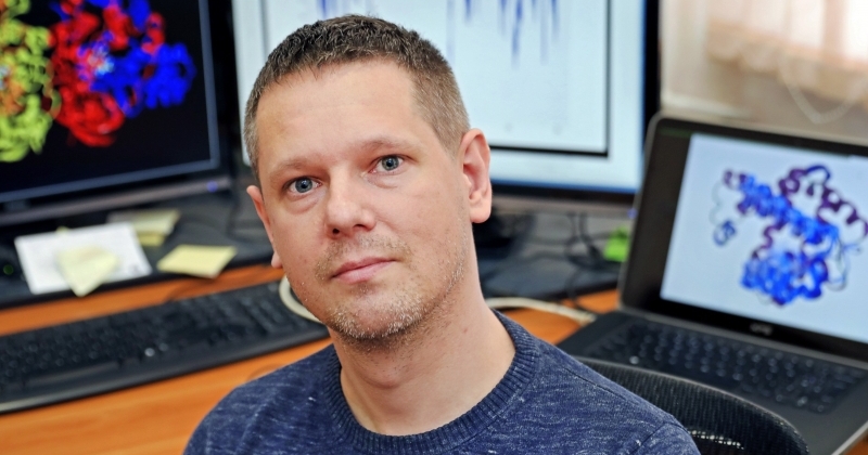 Portret dr. hab. Łukasza Pepłowskiego, prof. UMK, siedzącego przy biurku na tle monitorów wyświetlających trójwymiarowe modele struktur molekularnych oraz wykresy badawcze.