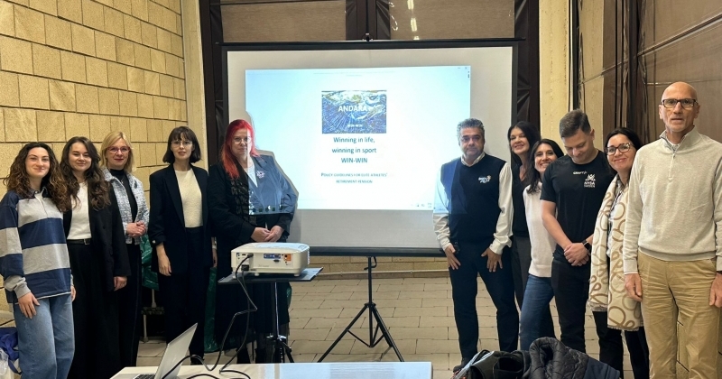 Grupowe zdjęcie uczestników międzynarodowego spotkania projektowego. Przedstawiciele partnerów stoją w sali konferencyjnej obok ekranu z prezentacją projektu ?Winning in life, winning in sport (WIN-WIN)?.