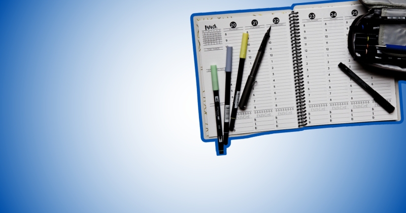 Na ciemnoniebieskim tle otwarty kalendarz książkowy. Na nim cztery kolorowe długopisy i czarny piórnik. 