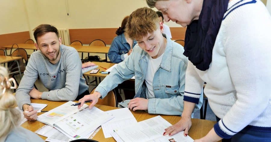Nauczycielka pochylona nad stołem pomaga dwóm uśmiechniętym studentom przeglądać materiały i notatki podczas wspólnej pracy w sali lekcyjnej.