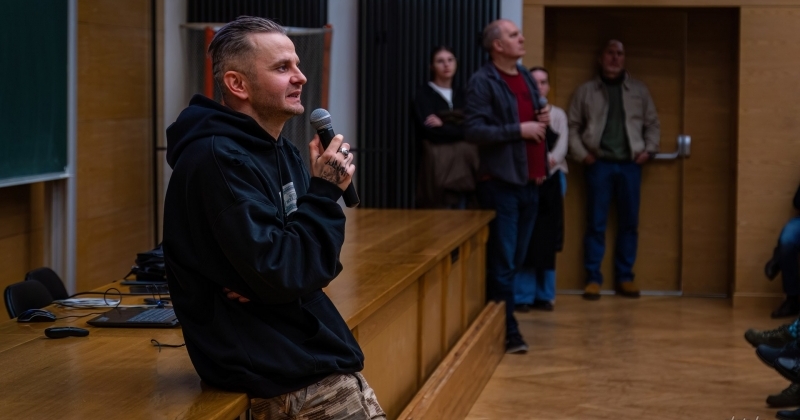 Prof. A. Dragan opiera się o biurko w Audytorium Aleksandra Jabłońskiegl, w tle widać trzy osoby