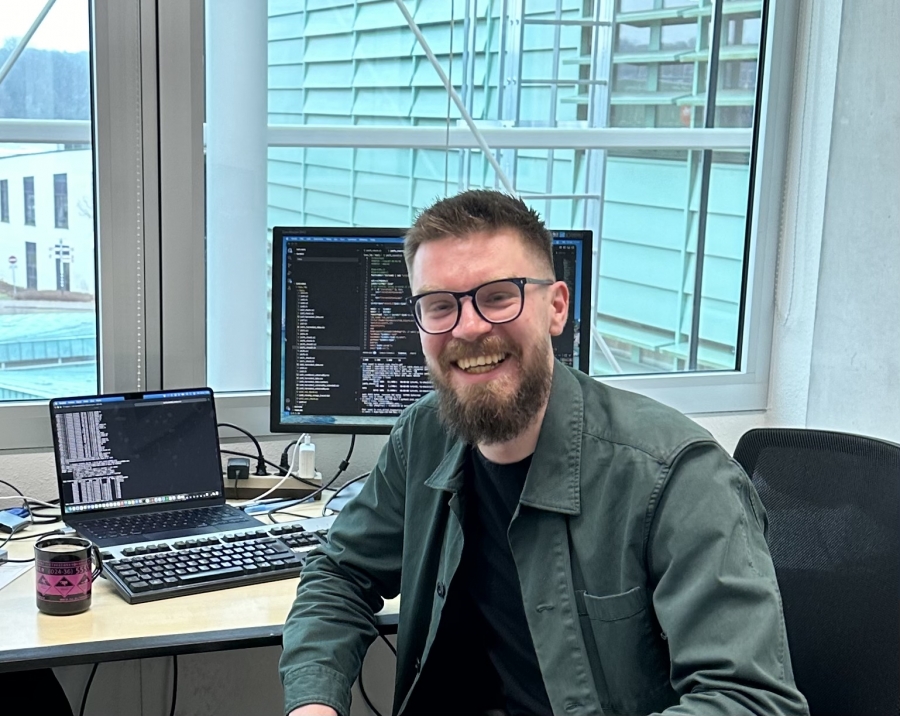 Uśmiechnięty mężczyzna z brodą i w okularach siedzi przy biurku w jasnym biurze. Na stanowisku pracy widoczny jest laptop oraz monitor z wyświetlonym kodem programistycznym.