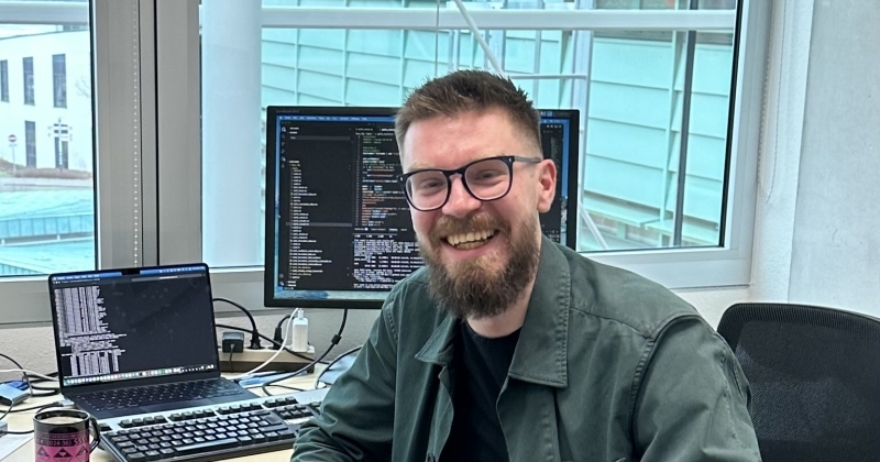 Uśmiechnięty mężczyzna z brodą i w okularach siedzi przy biurku w jasnym biurze. Na stanowisku pracy widoczny jest laptop oraz monitor z wyświetlonym kodem programistycznym.