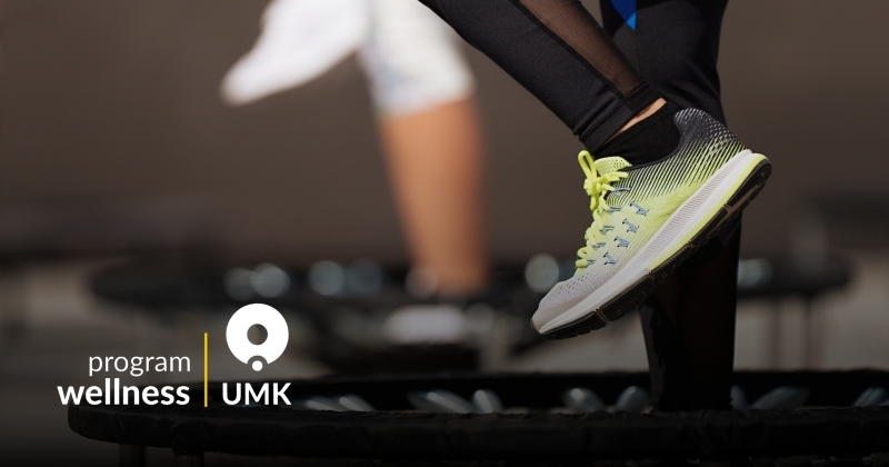 **Tekst alternatywny (alt):** Osoba ćwicząca na trampolinie podczas treningu fitness ? widoczne nogi w sportowych legginsach i butach, w tle rozmyta sala treningowa. Grafika z logo Program Wellness UMK.