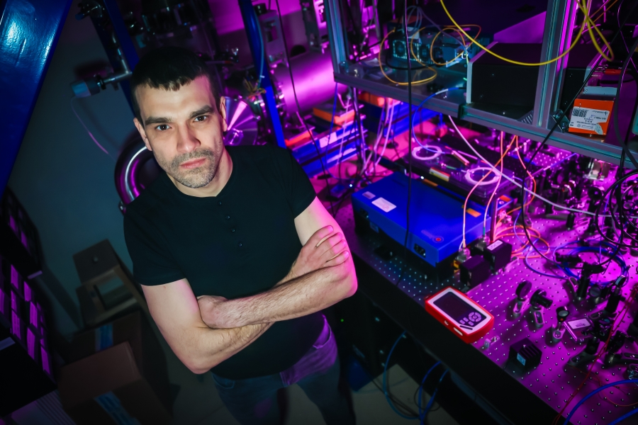 Na zdjęciu widoczny jest profesor Piotr Wcisło, mężczyzna o krótkich, ciemnych włosach, ubrany w czarny t-shirt i ciemne spodnie. Stoi z rękami skrzyżowanymi na piersi, patrząc bezpośrednio w stronę obiektywu z poważnym wyrazem twarzy. Zdjęcie zostało wykonane z góry (perspektywa wysoka), co pozwala objąć wzrokiem szeroki plan laboratorium. Tło i otoczenie wypełnia skomplikowana aparatura badawcza. Po prawej stronie widać stół optyczny z gęstą siecią luster, soczewek, niebieskich modułów elektronicznych oraz liczne kable (żółte światłowody i czarne przewody). Po lewej i w głębi znajdują się metalowe konstrukcje i elementy próżniowe. Całość jest skąpana w efektownym, niemal futurystycznym oświetleniu z dominacją barw magenty, fioletu i niebieskiego, które odbijają się od metalowych powierzchni sprzętu.