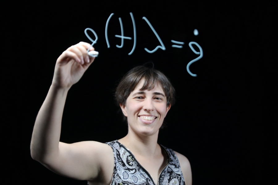 Na zdjęciu znajduje się uśmiechnięta dr hab. Katarzyna Siudzińska, prof. UMK, o krótkich, ciemnych włosach. Patrzy w stronę widza z entuzjazmem. Jest ubrana w ciemną bluzkę bez rękawów z jasnym, dekoracyjnym wzorem (paisley). Kobieta trzyma w prawej dłoni biały marker i pisze na niewidocznej, przezroczystej tafli szkła. Przed nią widnieje odręcznie napisany biały wzór matematyczny, który z perspektywy widza jest lustrzanym odbiciem (m.in. litery greckie, jak ? oraz symbole w nawiasach). Tło jest jednolicie czarne, co sprawia, że postać i białe napisy są bardzo wyraźne i mocno oświetlone. Światło pada z przodu, podkreślając twarz i dłoń piszącej osoby.