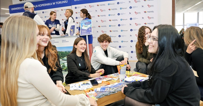 Grupa kandydatów i kandydatek na studia siedzą przy wspólnym stole, śmieją się i rozmawiają. Na stole leżą ulotki.