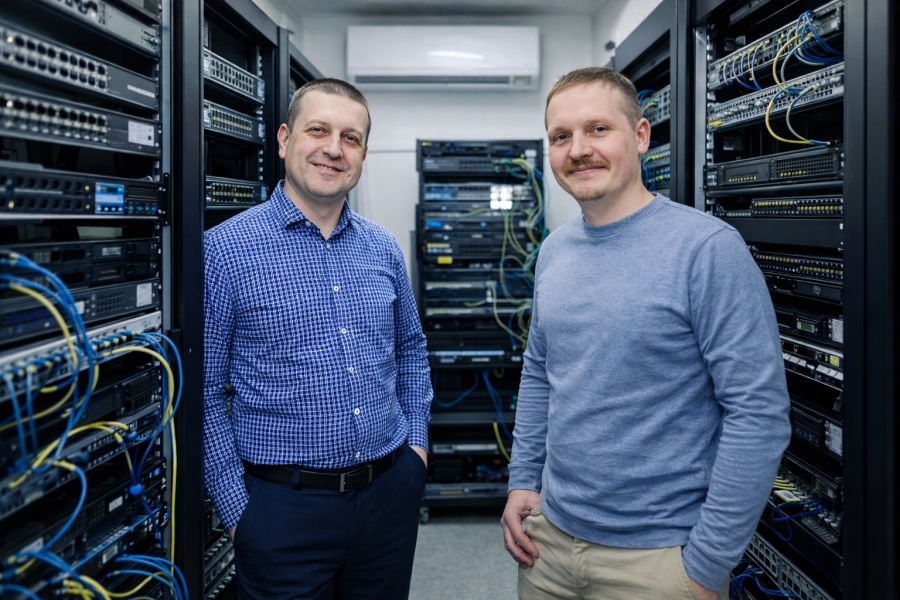 Two smiling IT specialists (prof. Piotr Masłowski and prof. Mariusz Piwiński) standing in a server room between racks of networking equipment.