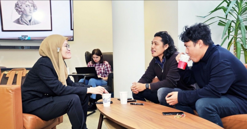 Three international students are sitting at a table drinking coffee; in the background, one person is working on a laptop, and a portrait of Nicolaus Copernicus can also be seen in the background