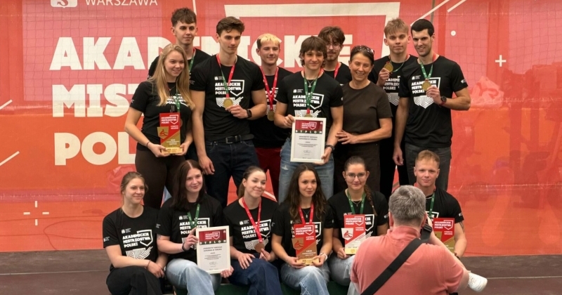 Grupowe zdjęcie reprezentacji UMK po Akademickich Mistrzostwach Polski w ergometrze wioślarskim. Zawodniczki i zawodnicy pozują z medalami, dyplomami i pucharami na tle czerwonej ścianki z napisem ?Akademickie Mistrzostwa Polski?. W centrum stoi także opiekunka grupy.