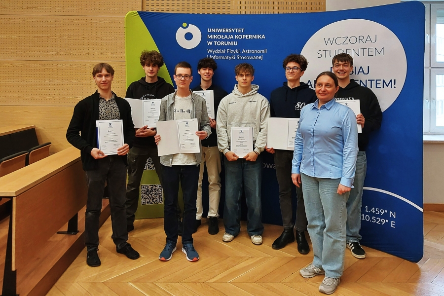 Osoby stojące i trzymające dyplomy w Audytorium Aleksandra Jabłońskiego na tle ścianki z logo WFAiIS