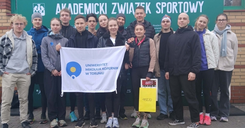 Grupowe zdjęcie studentów Uniwersytetu Mikołaja Kopernika w Toruniu, uczestników Akademickich Mistrzostw Polski. Grupa pozuje na tle zielonego baneru z napisem 