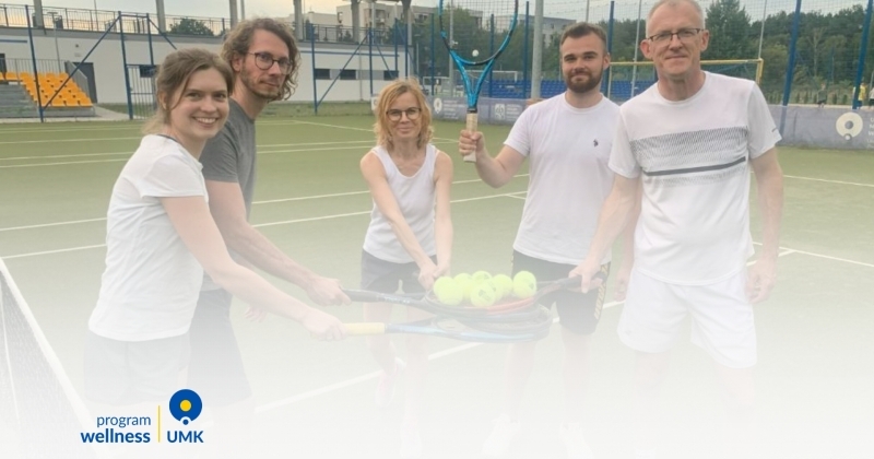 Grupowe zdjęcie pięciorga uczestników Programu Wellness UMK, pozujących na korcie tenisowym Uniwersyteckiego Centrum Sportowego. Grupa trzyma rakiety tenisowe, tworząc platformę z piłkami w centralnym punkcie zdjęcia. Wszyscy są ubrani w białe stroje sportowe. W tle widoczne są trybuny i infrastruktura obiektu, a w lewym dolnym rogu znajduje się logo Programu Wellness UMK.
