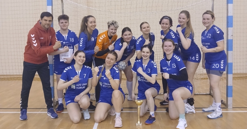 Drużyna piłki ręcznej kobiet AZS UMK Toruń pozuje do wspólnego zdjęcia w hali sportowej. Zawodniczki w niebieskich strojach oraz trener stoją i klęczą przed bramką, prezentując gest zaciśniętej pięści. Na środku, na parkiecie, ustawiony jest puchar.