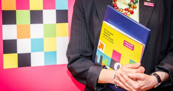 Zbliżenie na osobę trzymającą katalog z programem 24. Toruńskiego Festiwalu Nauki i Sztuki. W tle widać ściankę z logo Festiwalu