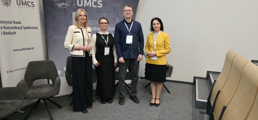 Cztery osoby stoją obok siebie podczas wydarzenia akademickiego, na tle banerów UMCS i Instytutu Nauk o Komunikacji Społecznej i Mediach; mają identyfikatory konferencyjne i są elegancko ubrane, a obok znajdują się krzesła oraz fragment sali wykładowej.