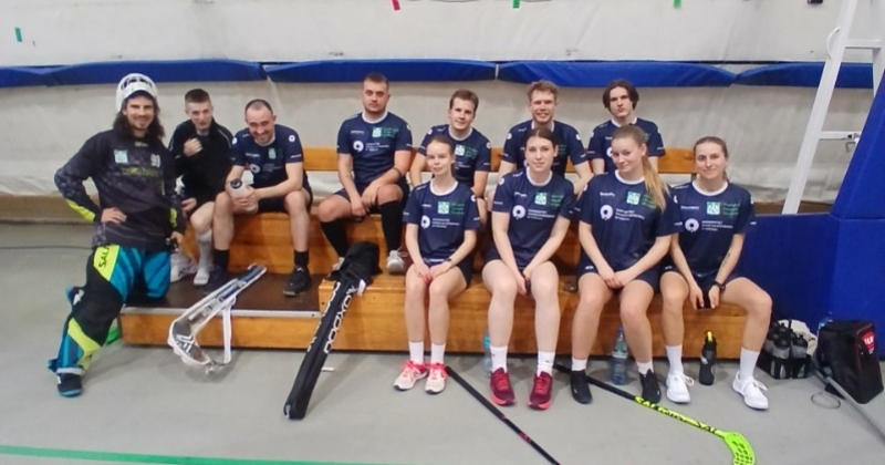 Mieszana drużyna unihokeja w granatowych strojach pozuje wspólnie z bramkarzem w hali sportowej, trzymając kije i sprzęt siedząc na drewnianych trybunach.