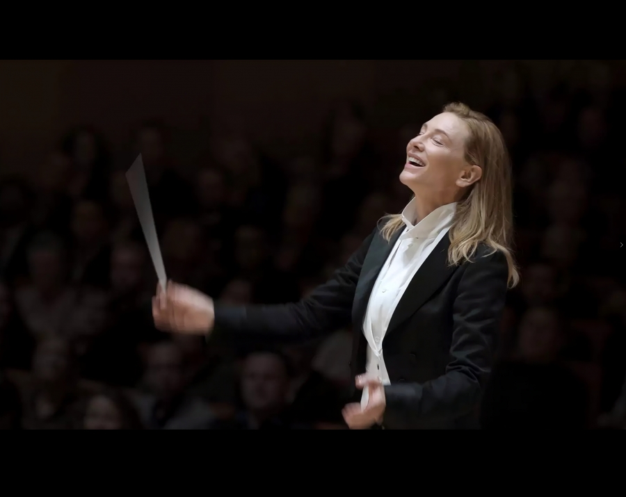 całość w czerni. Po środku kobieta w blond włosach rozpuszczonych do ramion. W czarnej marynarce i białej koszuli . Trzyma w prawej dłoni batutę. Widać, że bauta jest w ruchu. 