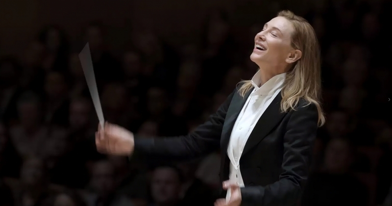 całość w czerni. Po środku kobieta w blond włosach rozpuszczonych do ramion. W czarnej marynarce i białej koszuli . Trzyma w prawej dłoni batutę. Widać, że bauta jest w ruchu. 
