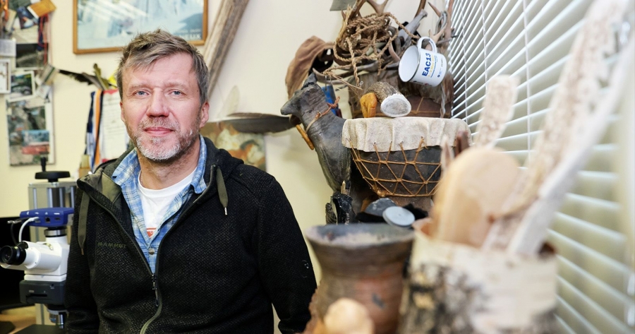 Osoba stojąca w pracowni archeologicznej obok mikroskopu, na tle półek z narzędziami i eksponatami z kości, poroża i ceramiki.