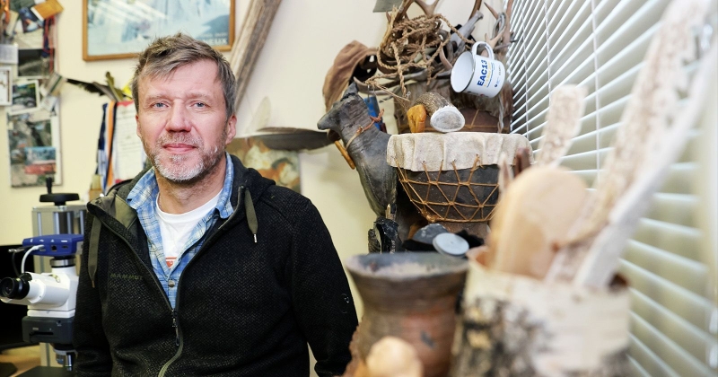 Osoba stojąca w pracowni archeologicznej obok mikroskopu, na tle półek z narzędziami i eksponatami z kości, poroża i ceramiki.